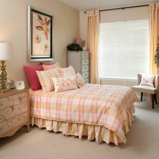 A cozy bedroom with a pink and beige color scheme featuring a neatly made bed, decorative pillows, a nightstand with a lamp, and a window dressed with light-colored curtains.