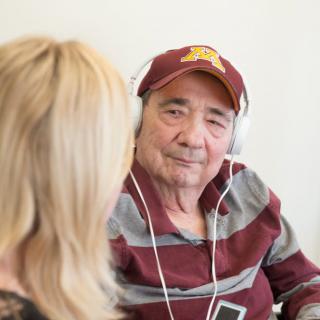An elderly man wearing headphones and a baseball cap engages in a conversation indoors.