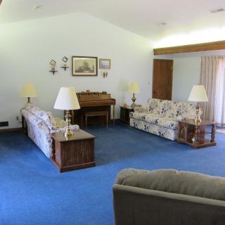 A vintage-style living room featuring blue carpeting, floral-patterned sofas, a piano, and multiple lamps.