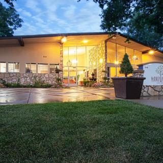 A modern building with illuminated windows during the evening, surrounded by well-maintained landscaping and trees.