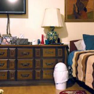 A cozy bedroom featuring a wooden dresser with a lamp and various items on top, a television mounted on the wall, and part of a bed with colorful pillows and a patterned blanket.