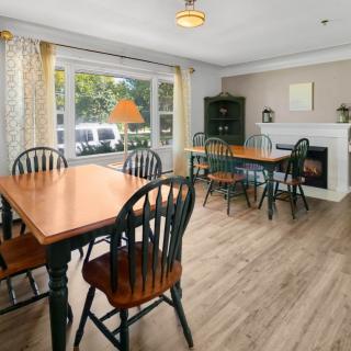 A cozy dining area with wooden tables and chairs, featuring a fireplace and large window with curtains.
