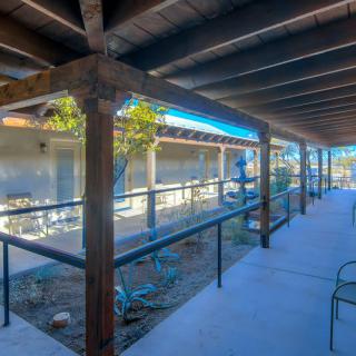 A sunny outdoor corridor with wooden beams and a ramp leading to an adjacent building.