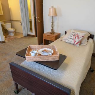 A cozy bedroom featuring a single hospital-style bed with a breakfast tray, a lamp on the nightstand, and an adjoining bathroom.