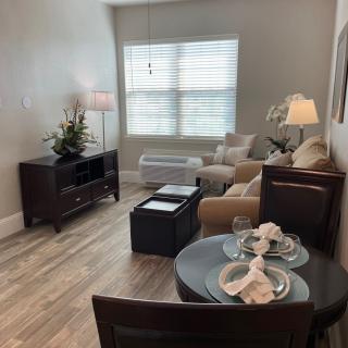 A cozy, modern living room with light wooden flooring, featuring a comfortable seating area and a small dining table near the window.