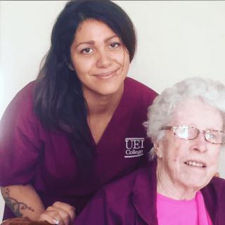 A caregiver in a nurse uniform poses with an elderly woman, both smiling.