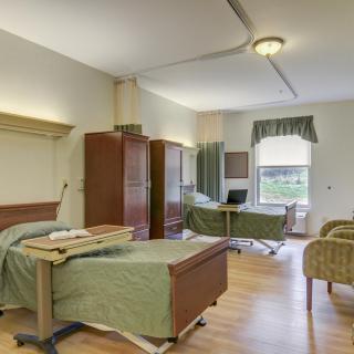 A hospital room featuring two beds, wardrobes, chairs, and a window with green curtains.