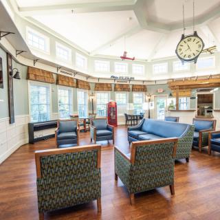 A spacious lounge with comfortable seating, large windows, and a prominent clock on the wall.