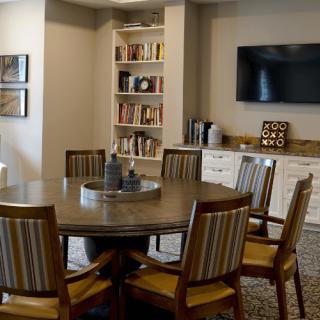 A cozy meeting room with a round table, surrounded by chairs and shelves filled with books, featuring a wall-mounted television.
