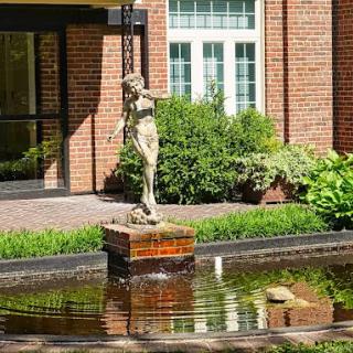 A decorative statue stands in the center of a garden fountain, set against a backdrop of lush greenery and a brick building.