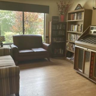 A cozy living room with comfortable seating, a jukebox, and bookshelves next to a large window.