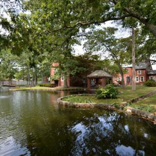 A tranquil scene of a brick building by a lakeside, surrounded by lush green trees and clear reflections in the water.