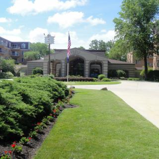 A landscaped entrance with a flag, surrounded by greenery and trees under a clear sky.