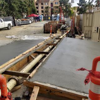 A view of a construction site where concrete has been recently poured, with visible safety barriers and plumbing elements.