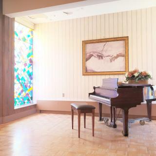 A serene room featuring a grand piano and stool, adorned with a painting of hands above. Vibrant stained glass and a pot of flowers add to the peaceful ambiance.