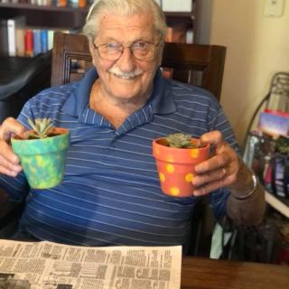 A smiling man holds two potted succulent plants indoors.
