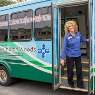 A smiling person stands at the open door of a bus, wearing a uniform.