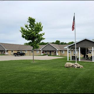 A view of single-story buildings with a neatly manicured lawn, a tree, and a flagpole under a cloudy sky.