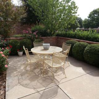 A cozy outdoor patio featuring a round table with four chairs, surrounded by lush greenery and neatly trimmed bushes.