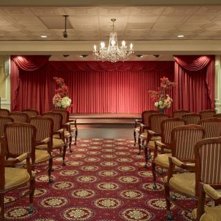 An elegant theater room with a stage set in front of red curtains, surrounded by rows of empty chairs and lit by a chandelier.