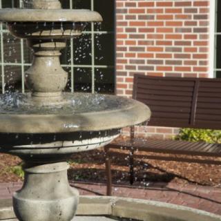 A stone fountain with flowing water next to a wooden bench, set against a brick wall outdoors.