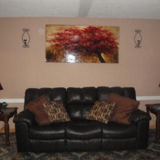 A cozy living room setup featuring a black leather sofa with decorative pillows, flanked by side tables and lamps. A vibrant painting of a tree adorns the wall above.