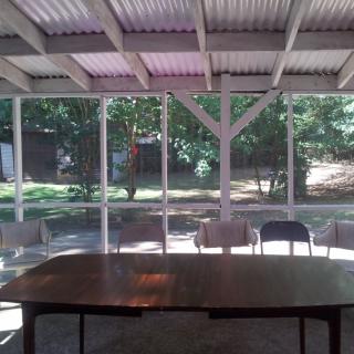 A covered outdoor area with a table and chairs, surrounded by trees and sunlight filtering through.