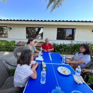 A cheerful family gathering around a blue table enjoying snacks on a sunny day.