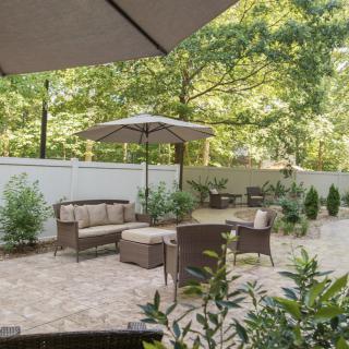 A peaceful patio with comfortable outdoor seating and an umbrella, surrounded by greenery and a white fence.