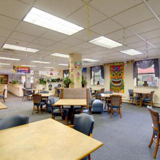 A spacious cafeteria with tables and chairs arranged for casual dining, featuring artwork on the walls and bright overhead lighting.