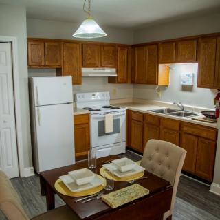 A cozy kitchen with wooden cabinets and a small dining area, featuring a refrigerator, stove, and a neatly set table.