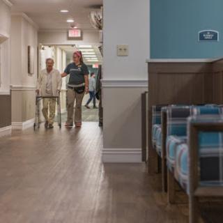A caregiver assists an elderly person in a well-lit hallway of an assisted living facility.