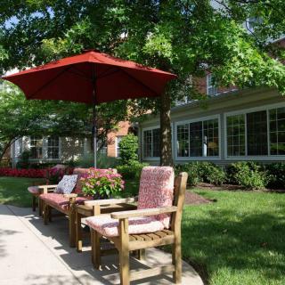 A serene outdoor patio area with wooden chairs and a red umbrella, surrounded by greenery.