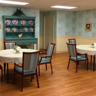 A cozy dining area featuring round tables with blue chairs, set on a wooden floor, complemented by floral wallpaper and a china cabinet.