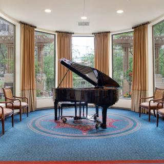 A grand piano in a bright music room with large windows and chairs.