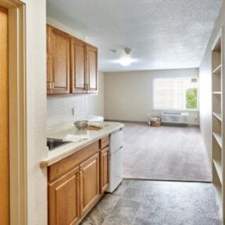 An empty apartment interior featuring a small kitchen area with wooden cabinets, leading into a bright living room with natural light.