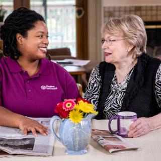 A caregiver engages in a warm conversation with an elderly woman, surrounded by flowers and morning coffee.