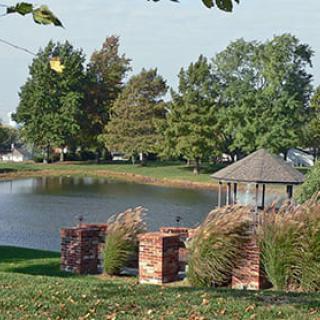 A serene landscape featuring a pond with a gazebo surrounded by lush trees.