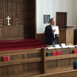 A person giving a presentation in a church auditorium, standing near the pulpit with a cross in the background.