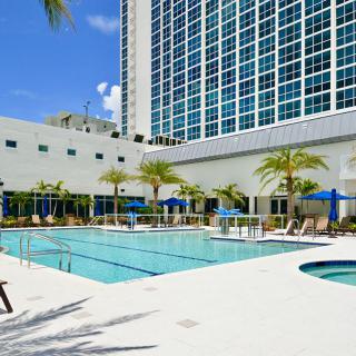 A modern hotel with a large outdoor swimming pool and palm trees on a sunny day.