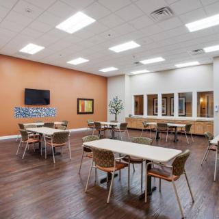 A modern cafeteria with wooden flooring, tables, and chairs arranged neatly under bright ceiling lights.