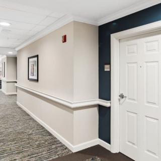 A well-lit hallway with a carpeted floor, featuring a closed door and framed artwork on the wall.