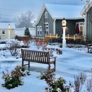 A snowy winter scene featuring a quaint house and garden with benches and a lamp post.