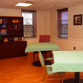 A bright room with tables and chairs, featuring a bookshelf and windows with blinds.
