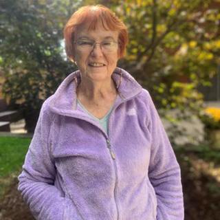 A woman wearing a purple jacket smiles while standing outdoors surrounded by greenery.