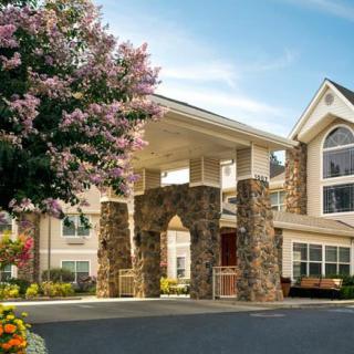 A picturesque building entrance with stone pillars, surrounded by trees and flowers under a clear sky.