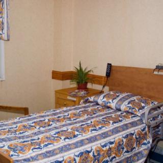 A simple bedroom with a single bed featuring patterned bedding, a plant on the nightstand, and a telephone on the wall.