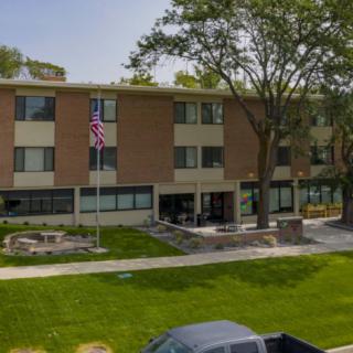 A multi-story brick building with a flagpole and well-maintained lawn, surrounded by trees and parked vehicles.