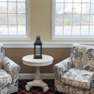A cozy living room corner with two patterned chairs flanking a round table topped by a lantern, set against large windows.