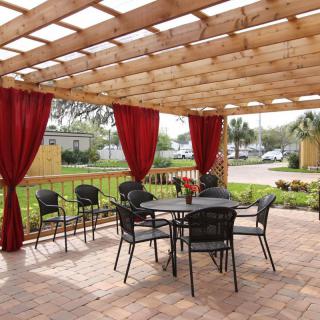A cozy outdoor patio with a wooden pergola and red curtains, featuring a table and chairs set on brick paving.